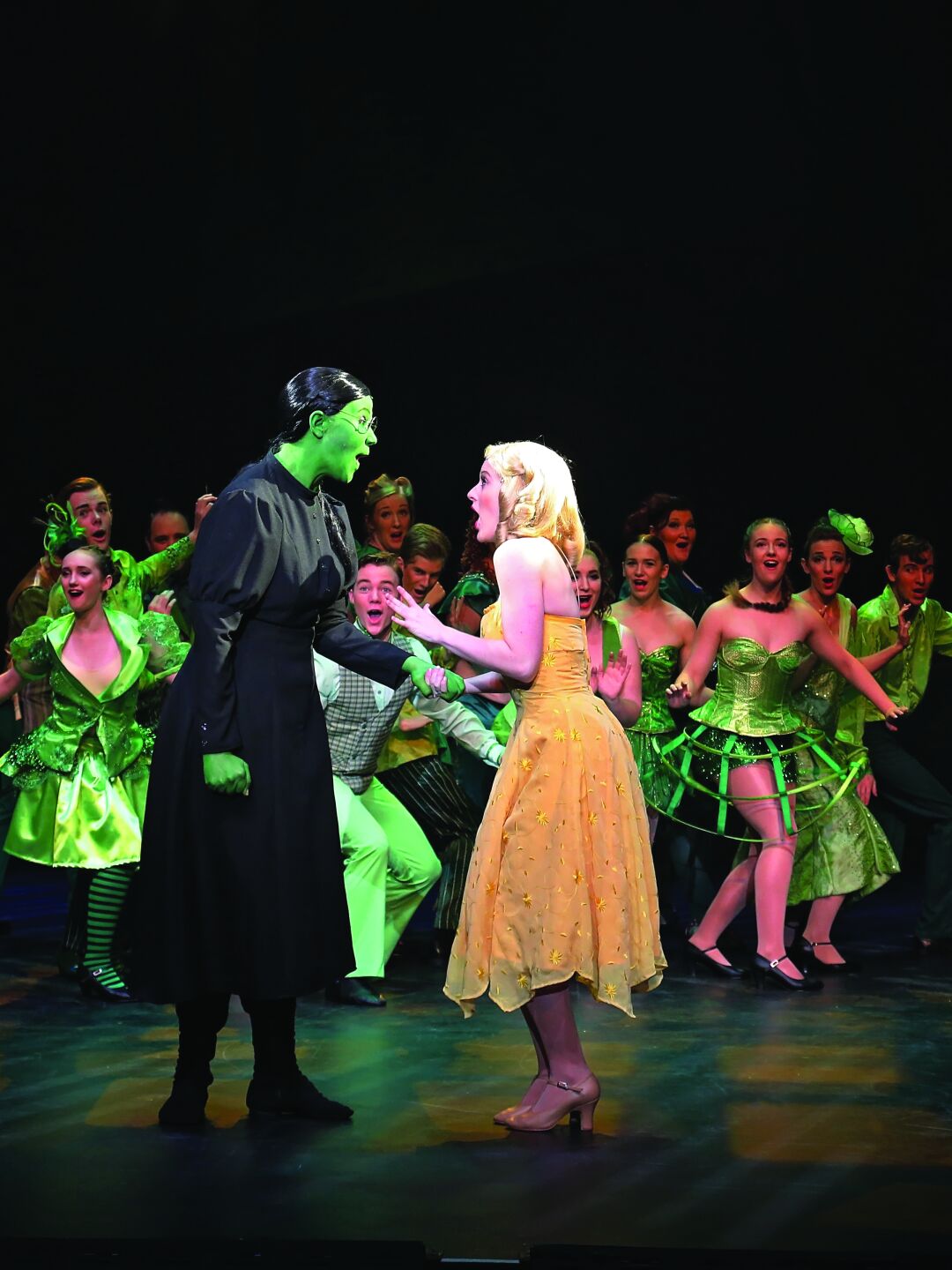 Two cast members in the forefront with large number of cast dancing in the background, during performance. 