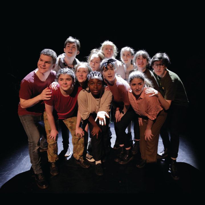 Group of young performers huddled together under a spotlight looking in different directions.